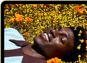 MacBook Neo, citrus color, screen shows a smiling person lying in a sunny field of flowers